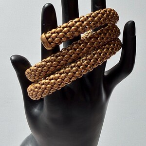 Könnte beinhalten: Goldfarbenes Armband mit strukturiertem, perlenbesetztem Design. Das Armband ist um eine schwarze, handförmige Präsentation gewickelt, die seine spiralförmige Form zeigt. Das Schmuckstück hat einen warmen, metallischen Glanz.