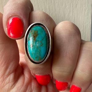 May include: A silver ring featuring a large, oval-shaped turquoise stone with brown matrix veining. The stone is set within a smooth, polished silver bezel that is framed by a secondary, slightly larger oval silver band, creating a layered effect.