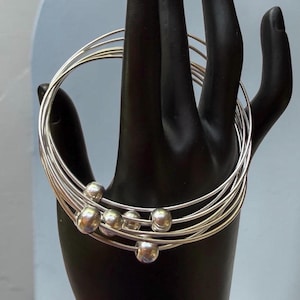 Könnte beinhalten: Ein Set silberfarbener Armreifen mit kleinen, runden Kugeln. Die Armreifen werden auf einer schwarzen, handförmigen Form vor einem hellblauen Hintergrund präsentiert. Die Armbänder bestehen aus dünnem, glänzendem Metall.
