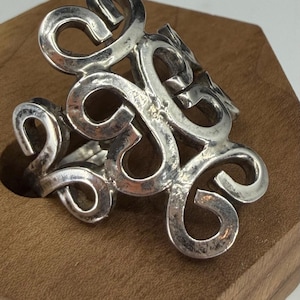 May include: A silver-colored ring with an openwork design of swirling, looping shapes. The ring is made of a shiny metal and sits on a wooden surface. The design is intricate and eye-catching, suitable for a fashion accessory.