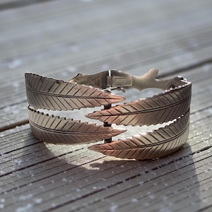 Könnte beinhalten: Silberfarbenes Armband mit Blattdesign. Das Armband hat drei Reihen stilisierter Blätter mit detaillierten Adern. Der Verschluss ist sichtbar, und es liegt auf einer Holzoberfläche.