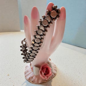 May include: A silver-toned bracelet with a repeating floral design is displayed on a pink ceramic hand. The bracelet's links feature small, detailed flower-like patterns. The hand is part of a decorative figurine with a rose at the base.