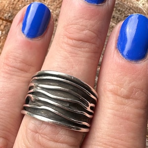 May include: A wide, silver-toned ring with a textured, wave-like design. The ring is worn on a finger with blue nail polish. The ring's surface has a dark, oxidized finish, enhancing the details of the flowing lines.