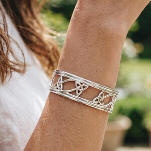 May include: A silver cuff bracelet with a Celtic knot design. The bracelet is rectangular and features a series of interconnected knot patterns. The metal has a polished, reflective surface. The bracelet is worn on an arm.
