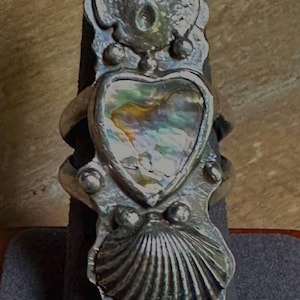 May include: A silver ring with a heart-shaped abalone shell inlay. The ring has a seashell design at the bottom and a skull-like design at the top. The ring is displayed on a grey ring holder.
