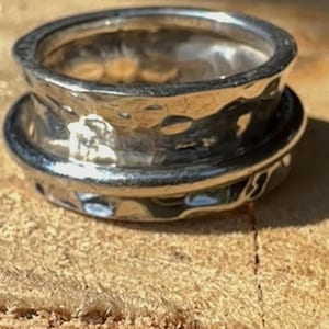 Peut inclure: Une bague argentée avec une texture martelée. La bague a une large bande avec une bande extérieure rotative. La bague est présentée sur une surface en bois.