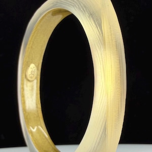 May include: A translucent, light yellow bangle bracelet with a textured surface. The interior is a warm gold colour. The bracelet has a rounded, oval shape and is set against a black background.
