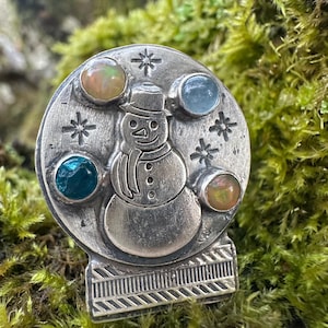 Puede incluir: Un amuleto de bola de nieve de plata con un muñeco de nieve y cuatro piedras preciosas de colores. El amuleto presenta un muñeco de nieve con un sombrero negro y una bufanda, y copos de nieve alrededor del borde.