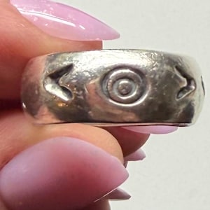May include: A silver ring with a wide band. The ring features a central circular design with concentric circles and stylized arrow-like shapes. The ring is held between fingers with pink nail polish.