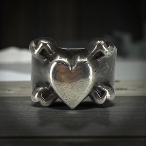May include: A silver ring with a heart-shaped design in the center. The ring has a textured surface and features crossbones on either side of the heart. The ring is a bold statement piece.