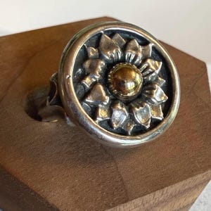 May include: A circular silver ring featuring a raised, multi-petaled floral design with a polished gold-tone center bead. The metal surface displays a darkened, oxidized finish within the recessed areas of the petals, creating a high-contrast look.