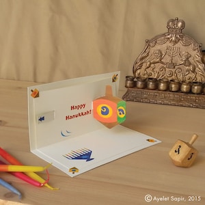 May include: A pop-up card with a colorful dreidel and the text "Happy Hanukkah!" The card also features a menorah with lit candles. A wooden dreidel is on the table next to the card.
