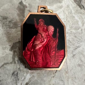 Könnte beinhalten: Ein achteckiger Metallanhänger mit einer roten und schwarzen Emaileinlage. Das Design zeigt eine Figur, die ein Kind auf den Schultern trägt und einen Stab hält. Der Anhänger hat einen goldfarbenen Metallrand und eine Öse an der Oberseite.