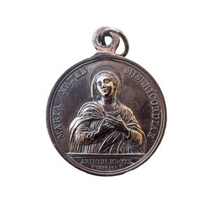 Puede incluir: Un colgante redondo de plata antigua con un relieve detallado de una mujer con halo, inscrito con las palabras "MARIA MATER MISERICORDIAE". El colgante tiene un lazo en la parte superior para colgar.