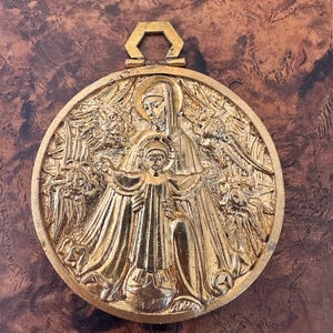 May include: A gold-colored, circular pendant featuring a detailed relief of a religious scene. The design includes figures of a woman and a child, surrounded by floral and angelic elements. The pendant has a hexagonal loop at the top for hanging.