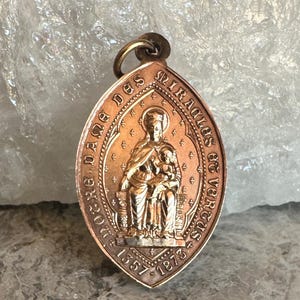 Könnte beinhalten: Ein kupferfarbener, ovaler Anhänger mit einer zentralen, geprägten Figur einer sitzenden Person, die ein Kind hält. Der Rand enthält den Text "ROGER DANE DES MIRACLES ET VERTUS" und die Jahreszahlen "1357-1873" mit kleinen Kreuzsymbolen.