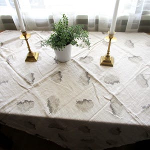 May include: A beige tablecloth with a floral pattern, two brass candle holders with white candles, and a small white pot with greenery. The tablecloth is draped over a wooden table.