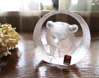 May include: A clear glass sculpture of a bear's face within a circular frame. The bear has detailed features and is set against a blurred background. A small, rectangular sticker is affixed to the base.