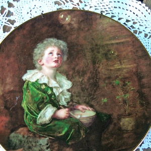 Op de afbeelding: Decoratieve plaat met een gouden rand, met een kind in een groen fluwelen pak met een witte gerimpelde kraag, die naar een bel kijkt. De achtergrond is donker en gestructureerd, met een kleine potplant aan de zijkant.