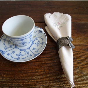 Op de afbeelding: Een wit theekopje met blauwe bloemenmotieven staat op een bijpassend schoteltje. Een witte servet met een bloemenborduursel wordt bij elkaar gehouden door een zilveren servetring.