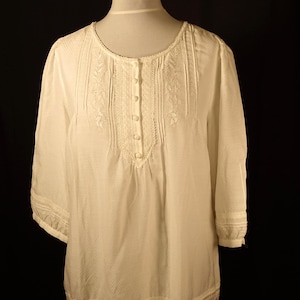 May include: White, long-sleeved blouse with a round neckline and button-down front. The blouse features intricate embroidered details on the bodice and sleeves, and a delicate lace trim at the hem. The fabric has a subtle dotted texture.
