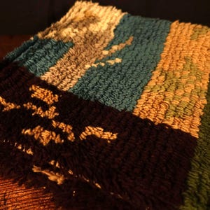 Op de afbeelding: Close-up van een veelkleurig tapijt met een getextureerd oppervlak. Het tapijt heeft strepen in bruin, blauwgroen, goud en groen. Het bruine gedeelte heeft een patroon van bruine vormen.