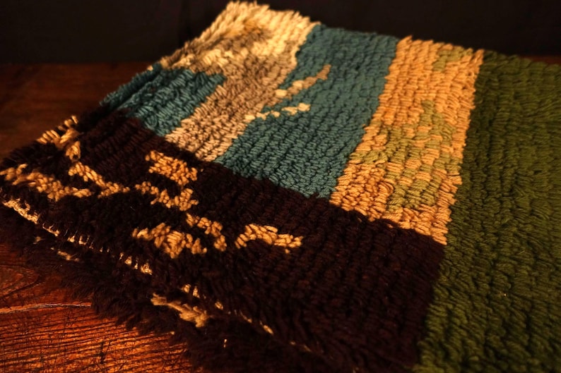 Op de afbeelding: Close-up van een veelkleurig tapijt met een getextureerd oppervlak. Het tapijt heeft strepen in bruin, blauwgroen, goud en groen. Het bruine gedeelte heeft een patroon van bruine vormen.