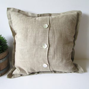 Peut inclure: Un coussin carré en lin beige avec trois gros boutons ronds nacrés au centre. Le coussin a un bord légèrement froissé et un aspect texturé. Une petite plante verte dans un tonneau en bois est visible en arrière-plan.
