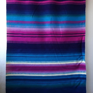 Op de afbeelding: Een levendig gekleurd textiel met horizontale strepen in tinten blauw, paars en roze. De stof heeft een gefranjerde onderrand en is opgehangen aan een metalen staaf met messingkleurige ringen. Het ontwerp doet denken aan een geweven deken of wandkleed.