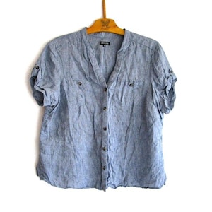 May include: A pale blue linen shirt with short sleeves, a V-neck, and a button-down front. The shirt features two small chest pockets and is suspended from a wooden hanger. The sleeves are rolled up, and the fabric has a textured finish.