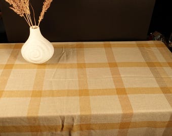 May include: A beige and yellow plaid tablecloth. A white vase with a spiral design holds dried wheat stalks. The tablecloth has a grid pattern with alternating beige and yellow stripes. The background is a dark brown.