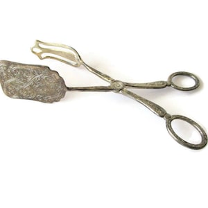 May include: A pair of silver-toned metal serving tongs featuring an ornate, embossed floral pattern on the rectangular gripping end and the handle. The tongs have two finger loops at the end and a hinged mechanism for opening and closing.