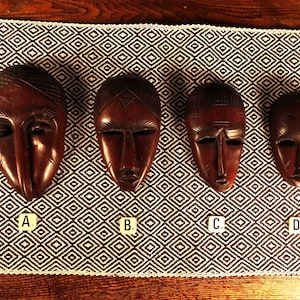 Op de afbeelding: Vier donkerbruine, uit hout gesneden maskers op een rij, gelabeld A, B, C en D. Elk masker heeft gestileerde gezichtsdetails, waaronder amandelvormige ogen, een prominente neus en gegraveerde haarpatronen, gerangschikt op afnemende grootte.