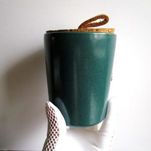 May include: A teal ceramic jar with a cork lid and a leather loop. The jar has a speckled texture and a cylindrical shape. The lid is made of cork and has a small leather loop attached for easy removal.