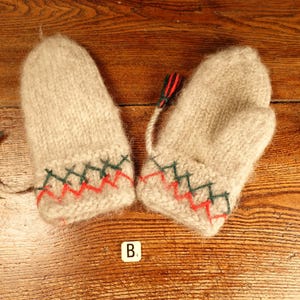 May include: A pair of beige fuzzy knit mittens featuring a decorative cuff with embroidered green and red zigzag stitching. Each mitten includes a small braided cord with a red and black tassel attached to the side of the wrist area.