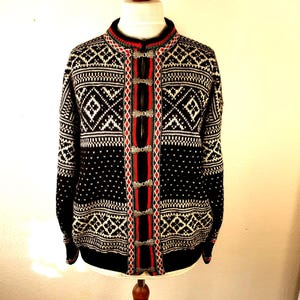 May include: A black knit cardigan featuring a white geometric diamond pattern and red accents along the front placket. The garment includes silver-toned metal clasps down the center, a high neckline, and long sleeves with matching intricate knit detailing.