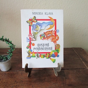 Pode incluir: Um livro infantil intitulado "Mirdza Klava" com ilustrações coloridas de um gato, um rato e outras imagens lúdicas. O livro é exibido em um suporte de latão, com uma pequena planta em vaso ao fundo.