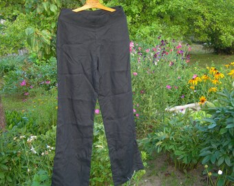 May include: A pair of solid black pants featuring a wide, flat waistband and a straight-leg cut. The pants are suspended from a wooden hanger, showcasing a smooth, matte fabric texture throughout the garment from the waist down to the hem.