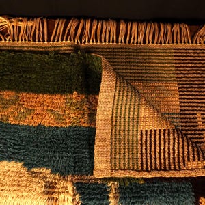 Op de afbeelding: Een kleurrijk, rechthoekig tapijt met een getextureerde, harige pool. Het tapijt heeft horizontale strepen in groen, goud en blauwgroen, met een gedeelte met verticale strepen in bruin en groen. Het tapijt heeft aan &eacute;&eacute;n kant franjes.