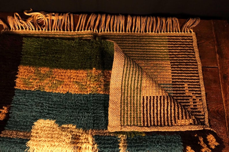 Op de afbeelding: Een kleurrijk, rechthoekig tapijt met een getextureerde, harige pool. Het tapijt heeft horizontale strepen in groen, goud en blauwgroen, met een gedeelte met verticale strepen in bruin en groen. Het tapijt heeft aan &eacute;&eacute;n kant franjes.