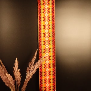 Könnte beinhalten: Ein langes, rechteckiges Textil mit einem geometrischen Muster in Orange-, Gelb- und Rottönen hängt vor einem dunklen Hintergrund. Das Textil hat oben und unten ein dekoratives Metallstück. Eine Vase mit getrockneten Pflanzen ist im Vordergrund.