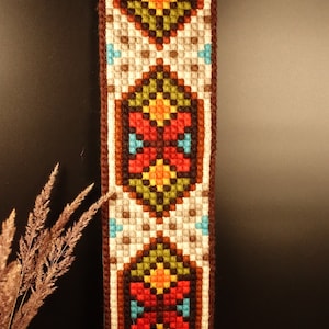 Könnte beinhalten: Vertikal dargestelltes gewebtes Textil mit einem sich wiederholenden geometrischen Muster in Rot, Orange, Gelb, Blau und Grün auf cremefarbenem Hintergrund. Die Ränder sind braun eingefasst. Der Hintergrund ist dunkel.