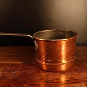 Op de afbeelding: Een koperen steelpan met een lange, zilveren handgreep. De pot heeft een afgeronde bodem en een cilindrisch lichaam. Het koper heeft een warme, roodbruine kleur en de handgreep is aan de zijkant bevestigd. De steelpan staat op een houten oppervlak.