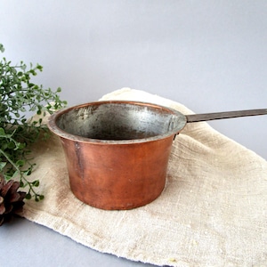 Peut inclure: Une casserole en cuivre vintage avec une longue poignée plate en métal. Le pot a un corps arrondi et un intérieur terni. Il repose sur un tissu beige, avec de la verdure et une pomme de pin en arrière-plan.