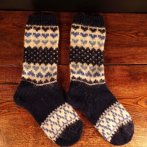 May include: A pair of navy blue knitted socks with a pattern of light blue and off-white hearts and diamonds. The socks are displayed on a wooden surface, showcasing their intricate design and cozy texture.