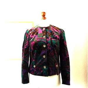 Puede incluir: Chaqueta de terciopelo vintage con un diseño vibrante y estampado en tonos morado, verde y rosa. La chaqueta presenta un cuello redondo, mangas largas y botones plateados. La tela tiene un aspecto suave y texturizado, y el estilo general recuerda a los años 80.
