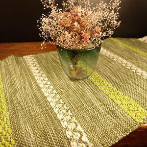 May include: A green woven table runner with white and yellow accents. A glass vase with dried flowers sits on the runner. The runner has a fringed edge and is displayed on a wooden surface.