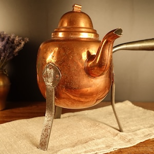 Puede incluir: Una tetera de cobre antigua con un pico y un asa, apoyada en un soporte de tres patas. La tetera tiene una tapa y está colocada sobre un paño de lino. Un jarrón de cobre con lavanda está en el fondo.