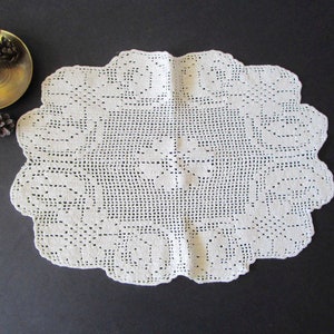 May include: Cream-coloured, oval-shaped crocheted doily with a scalloped edge. The doily features a repeating floral pattern. A brass candlestick and pine cones are visible in the background.