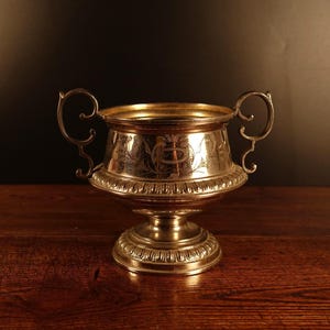 Könnte beinhalten: Ein silberfarbener Metall-Pokal mit zwei verzierten, geschwungenen Henkeln an den Seiten. Die Schale des Pokals ist mit filigranen Blumengravuren und einem horizontalen Band aus vertikalen Rillen über dem runden Sockelfuß verziert.